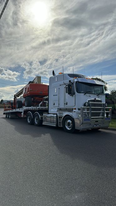 White semi-truck hauling heavy machinery on a sunny day with partly cloudy sky.