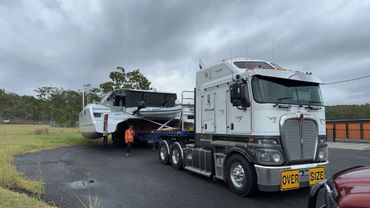 A large truck transporting a boat on a trailer with a person walking nearby.