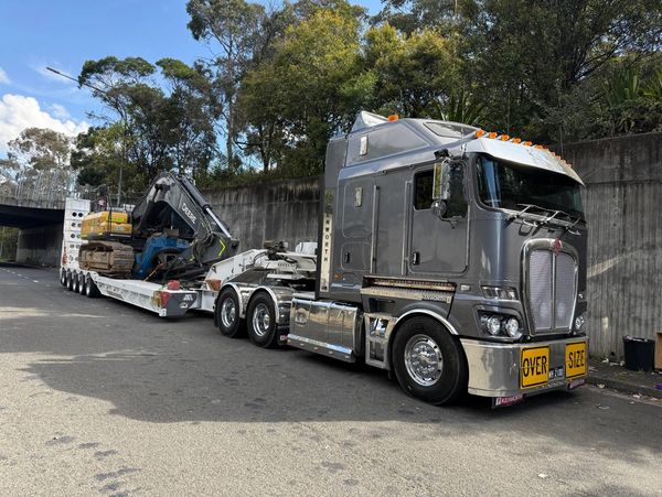 A silver Kenworth truck hauling a large excavator on a trailer in Newcastle