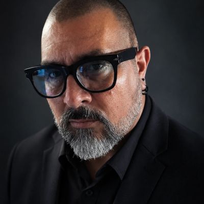 Serious man with glasses and beard in black suit.