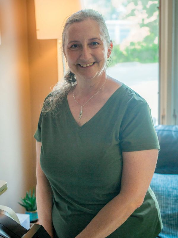 Smiling woman in green shirt standing indoors near a lamp and window.