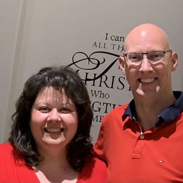 Smiling couple wearing red shirts standing indoors with a decorative wall quote behind them.