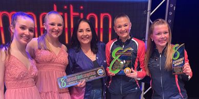 Group of smiling dancers and a woman holding dance competition awards and plaques.