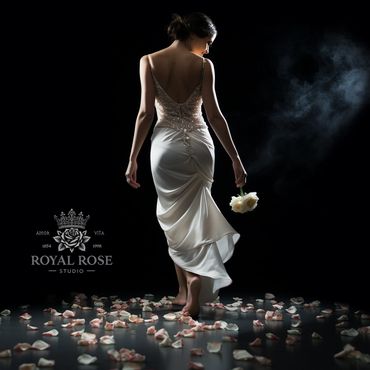 Woman in an elegant white gown walking barefoot on rose petals with a dark smoky background.