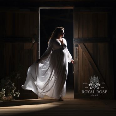 Pregnant woman in a flowing white dress standing in a doorway with dramatic lighting.