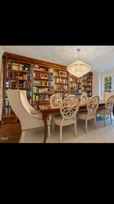 Elegant dining room with cream chairs, wooden table, large bookshelf, and chandelier.