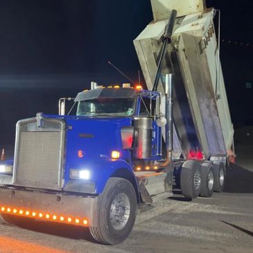 Blue dump truck unloading gravel at night with lights on.