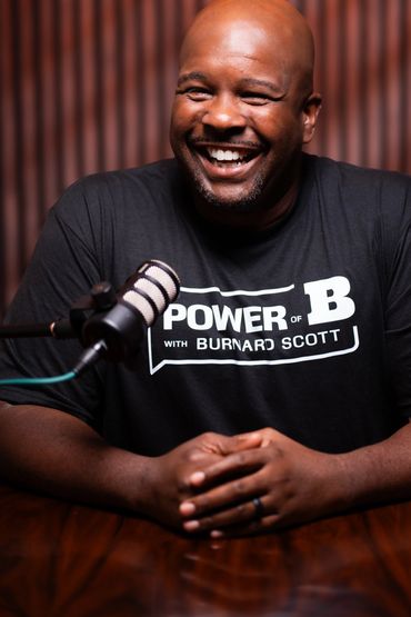 A smiling man in a black t-shirt sits behind a microphone.
