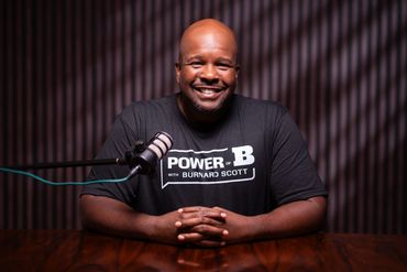 A smiling man sits at a table with a microphone, wearing a 'Power of B' t-shirt.