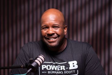 Smiling man wearing a "Power of B with Burnard Scott" t-shirt sitting at a table with a microphone.