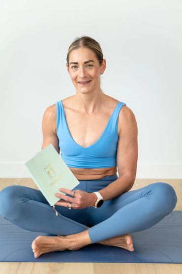 Woman in blue workout clothes sitting cross-legged on a mat, holding a book.