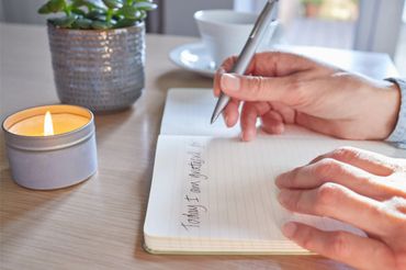 Person writing gratitude journal by candlelight with a plant and cup nearby.