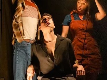 Four people in a dimly lit room, one holding a light bulb above another seated person.