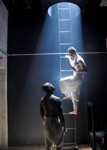 Two shirtless men in white pants by a ladder under a spotlight in a dark room.
