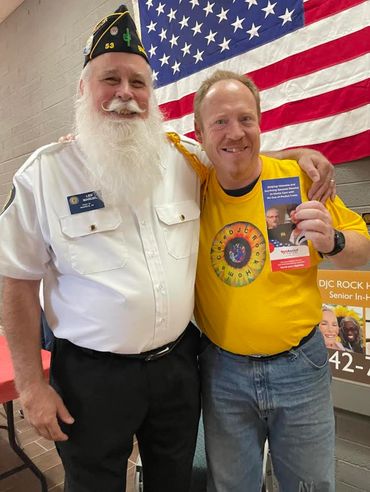 General manager in Company T shirt takes happy photo with senior Veteran in uniform with beard