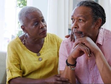 Skeptical black senior couple sits together as the wife reassures her husband