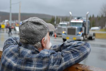 An elderly man with snow on his hat watches trucks on a rainy day.