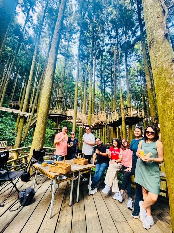 A group of friends enjoying drinks on a wooden deck in a forest with treehouses.