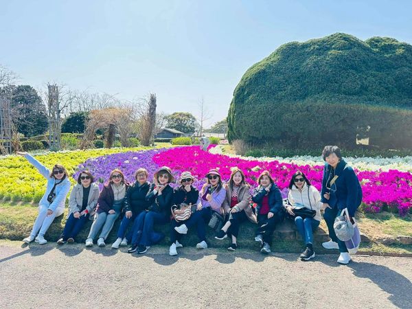 Group of women posing happily in front of colorful flower beds on a sunny day.
