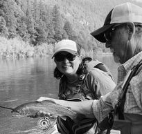A man holds a fish while standing on a boat in a river. A woman stands behind him smiling.