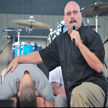 A man with glasses holds a microphone and places his hand on another man bending forward.