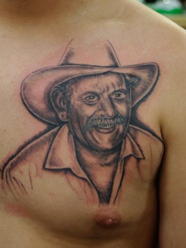 Black and grey portrait tattoo of an older man with a hat on a man's chest.