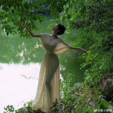 A woman in a flowing dress gracefully poses by a serene lakeside surrounded by lush greenery.
