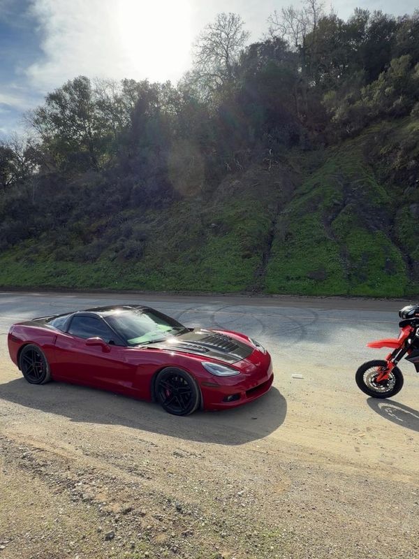 Red sports car and motorcycle parked on a sunny roadside near a green hillside.