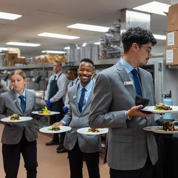 Waitstaff in gray suits carrying plated gourmet dishes in a restaurant kitchen.