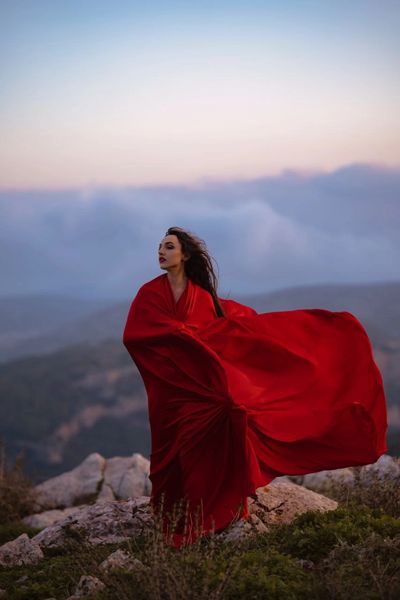 Woman in a flowing red dress stands on rocky terrain at dusk.
