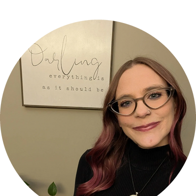 Woman with glasses and purple hair smiling in front of a motivational wall art.