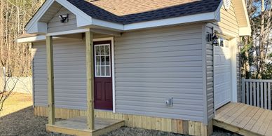 A small gray shed with a maroon door and a wooden ramp in a backyard.