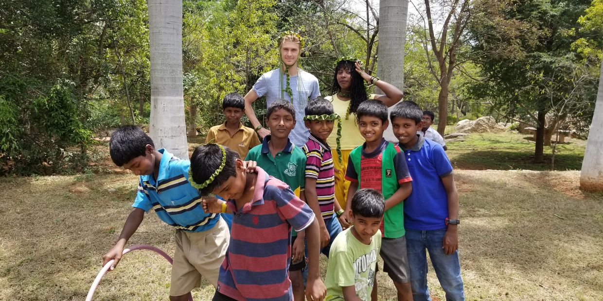 H.E. Cairo Eubanks wearing a palm tree crown surrounded by Indian youth who are posing with her and a fellow teacher. She is holding the crown on her head.