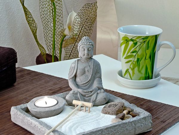 a Buddha, candle and sand in a concrete tray sat atop a wooden table. a green plant is in background