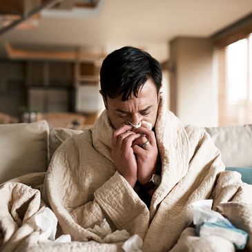 Man wrapped in a blanket, blowing his nose on a tissue.