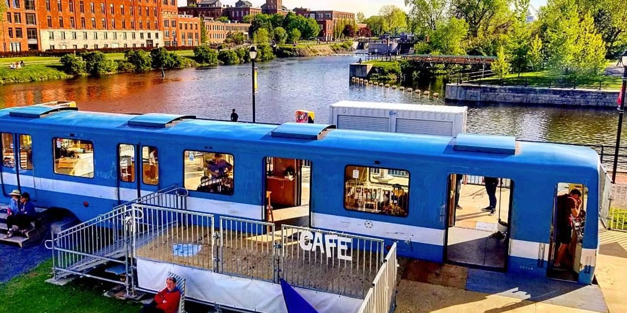 A blue train car converted into a café by a riverside with people inside and outside.