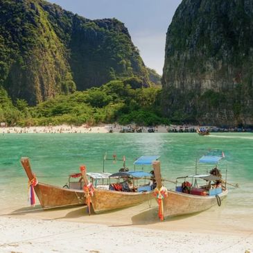 Three traditional wooden boats docked on a tropical beach with turquoise water and rocky cliffs.