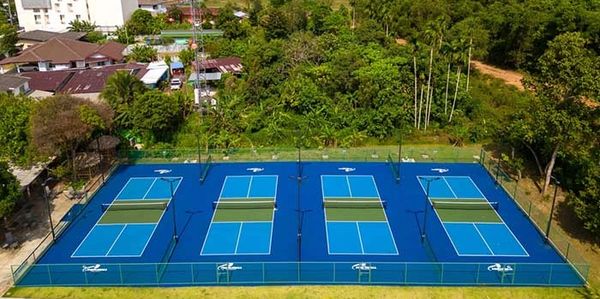 Aerial view of four blue tennis courts surrounded by greenery and buildings.