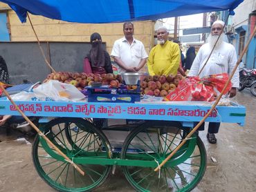 Four people standing behind a fruit cart with apples and a weighing scale.