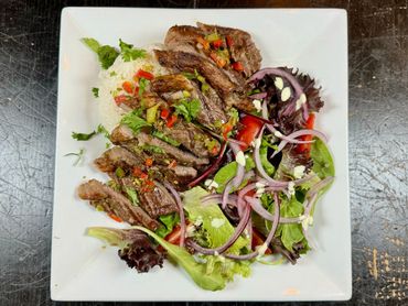 Grilled steak with mixed greens, red onions, and a side of white rice on a square plate.