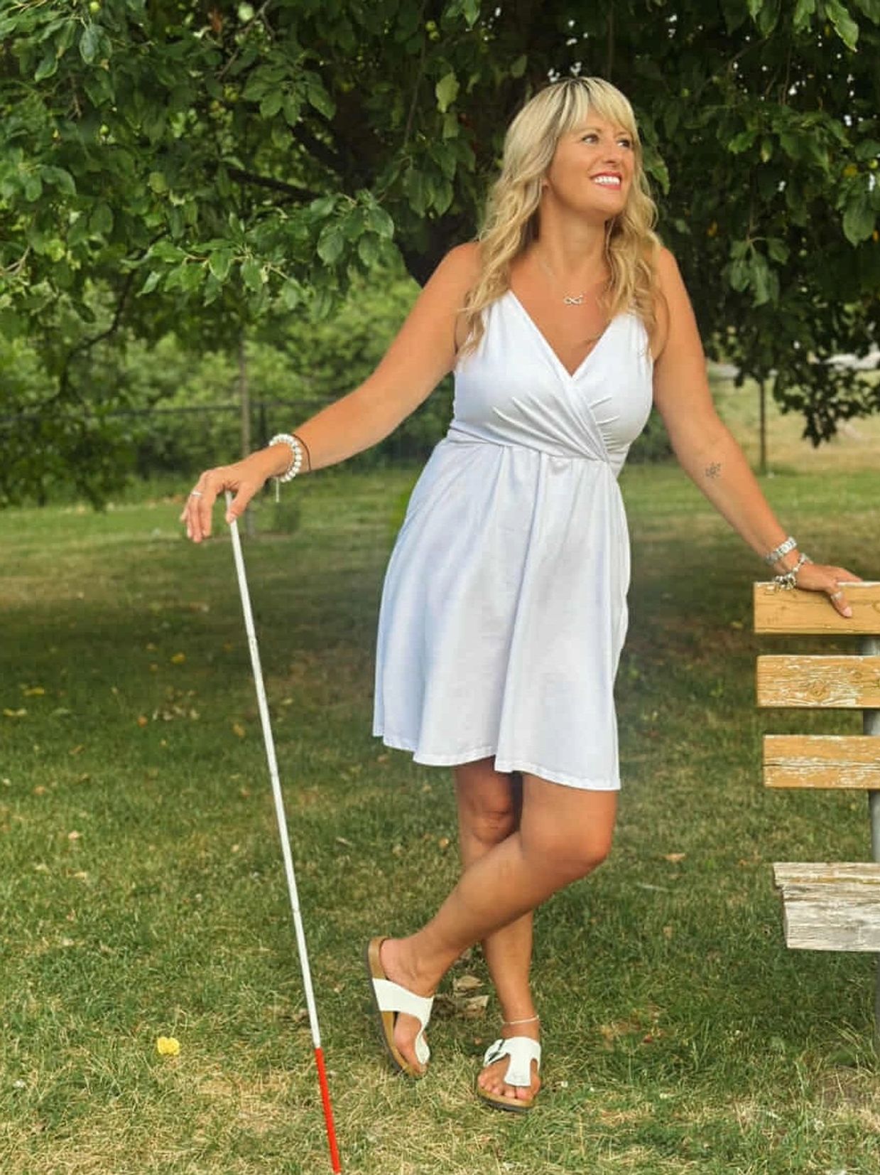 Smiling woman in white dress with white cane stands near a bench in a park.