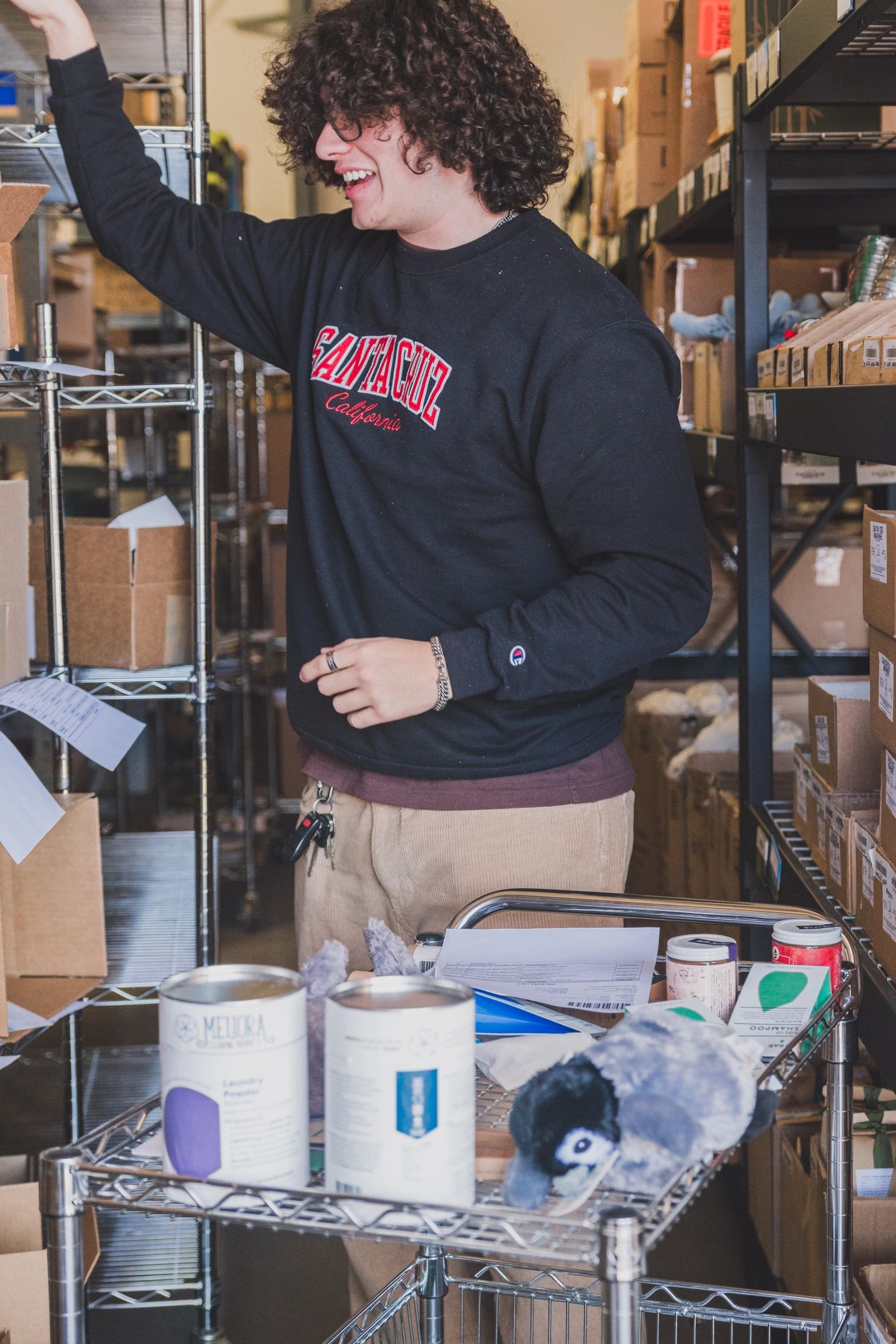 A worker in a black sweatshirt sorts products into boxes, surrounded by shelves.