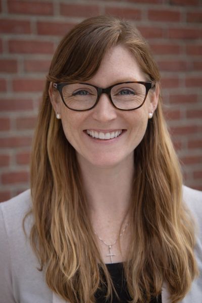 Smiling woman with glasses and long hair posing against a brick wall.