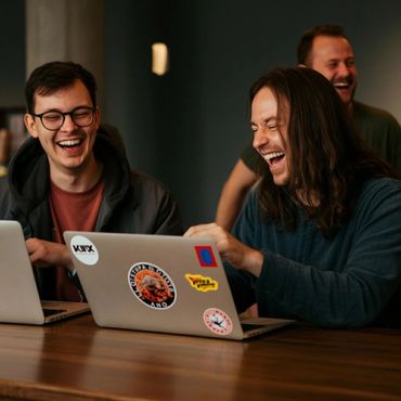 Three friends laughing together while using laptops at a wooden table.