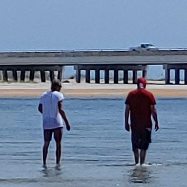 Two people walking in shallow water near a bridge.