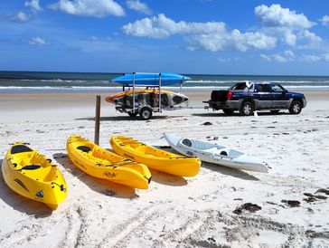 Yellow kayaks on a sandy beach near a blue truck and trailer.