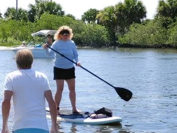 A woman paddleboarding on a calm river with a man watching from the shore.