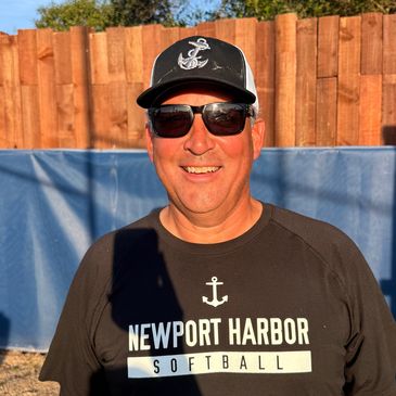 Man in sunglasses and hat smiling outdoors near a wooden fence.