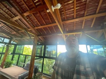 Man taking selfie in a sunlit wooden room with large windows and greenery outside.