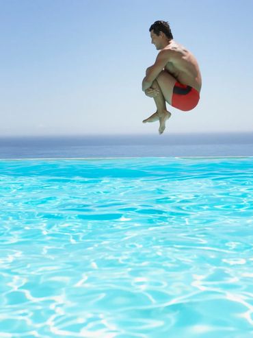 Man in red swim trunks jumping into a bright blue pool with ocean in background.